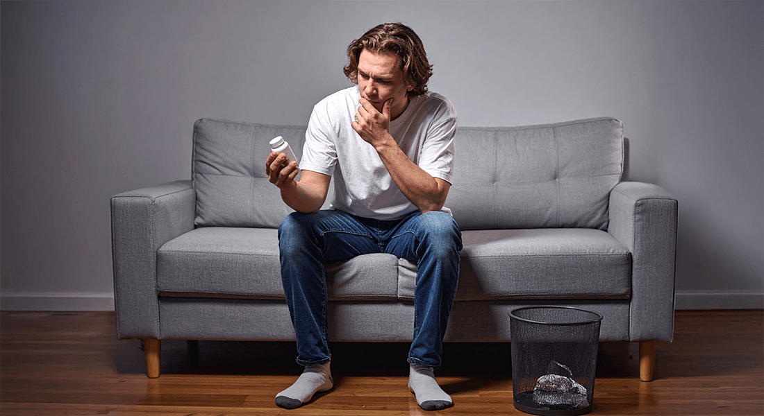 An image of a man on a sofa holding a bottle of supplements and wondering whether to throw them away. An image of a man on a sofa holding a bottle of supplements and wondering whether to throw them away.