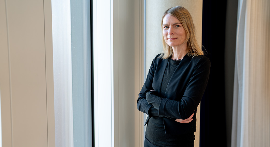Portrait photo of Camilla Schéele standing next to a window with her arms crossed and smiling at the camera
